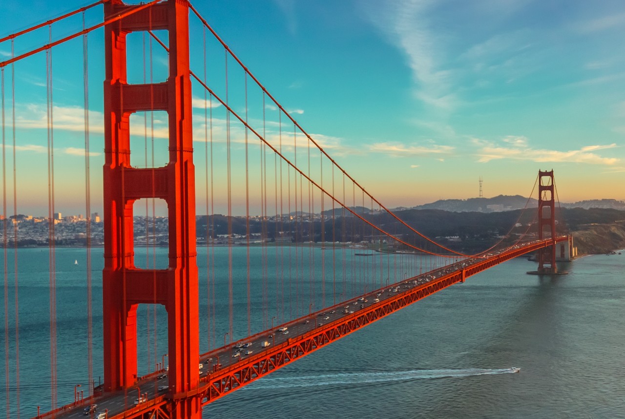Photograph of Golden Gate Bridge shortly after sunset. This is panoramic view of Golden Gate. Bridge is brightly illuminated and located on left side of photograph with colorful sunset sky.